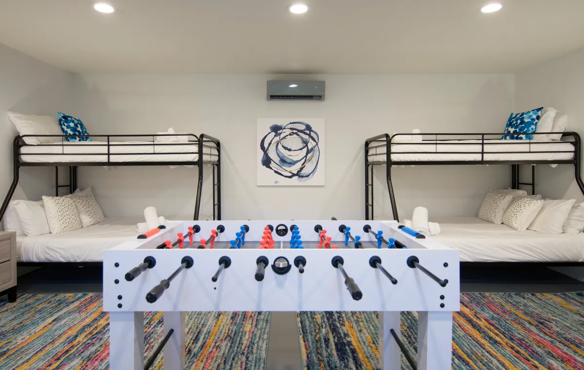 A fun, family-friendly room that features colorful bunk beds, a foosball table for play, a vibrant rug, and abstract wall art. The scene is lively, suggesting activities as well as a space for children or group sleepovers.
