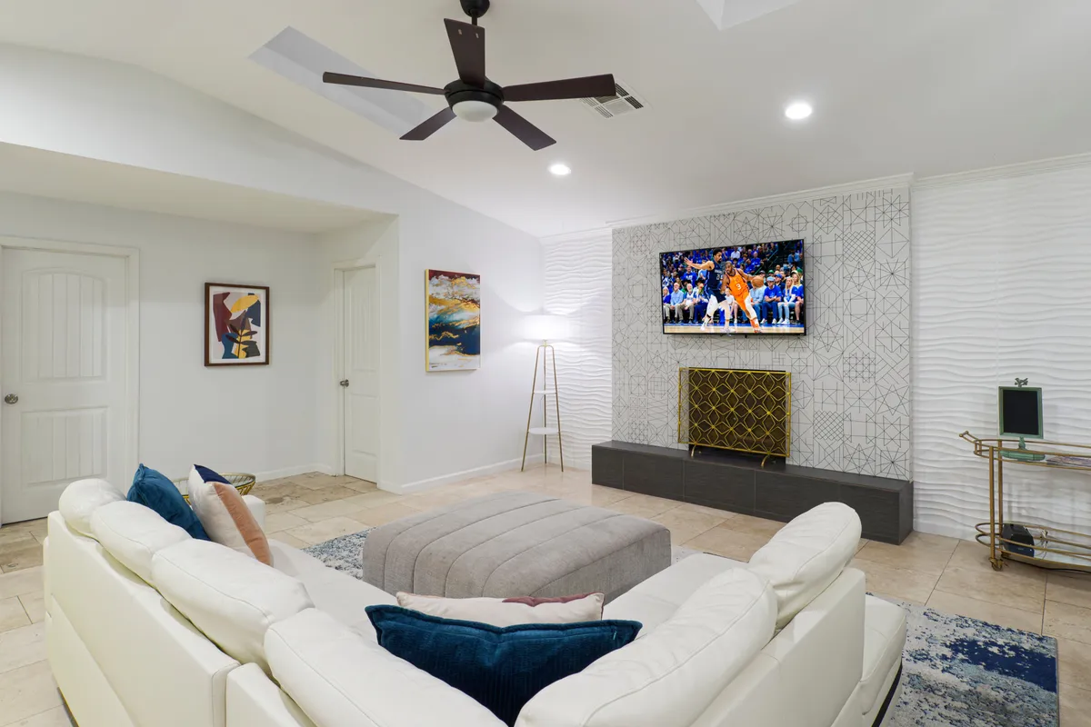 Showcasing a white sectional sofa facing a television with a sports game on, this interior scene also features textured wall decor, a floor lamp, and vibrant artwork. The mood is lively and comfortable, ideal for group entertainment and relaxation.