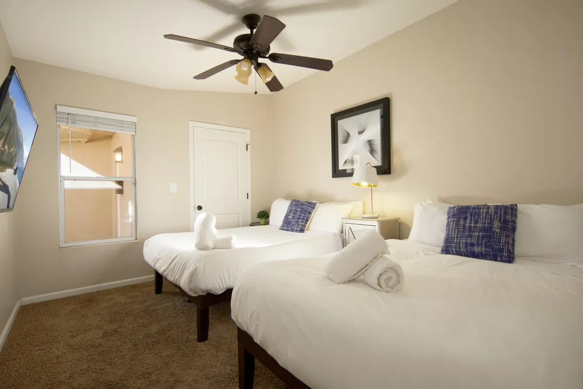 A comfortable bedroom setup with two beds featuring white bedding, wall art above, a ceiling fan, and a window letting in natural light. The room exudes a feeling of shared comfort, perfect for families or friends.