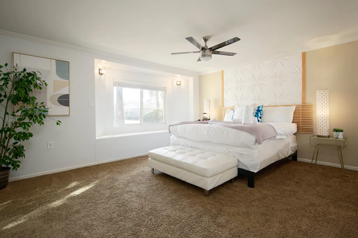 A serene bedroom featuring a plush bed with crisp bedding, a window allowing in natural light, decorative wall art, a plant on the bedside, and a nightstand with a lamp. The space feels both restful and stylish, inviting relaxation and comfort.