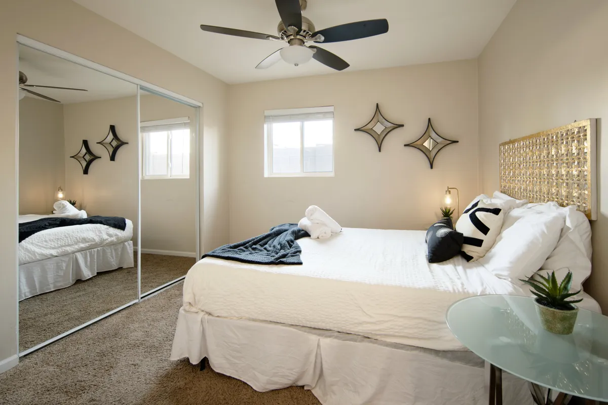 A cozy bedroom with a bed adorned with decorative pillows, a mirrored wardrobe, decorative wall mirrors, and a small table with a plant. Natural light fills the room, creating a serene setting ideal for relaxation.