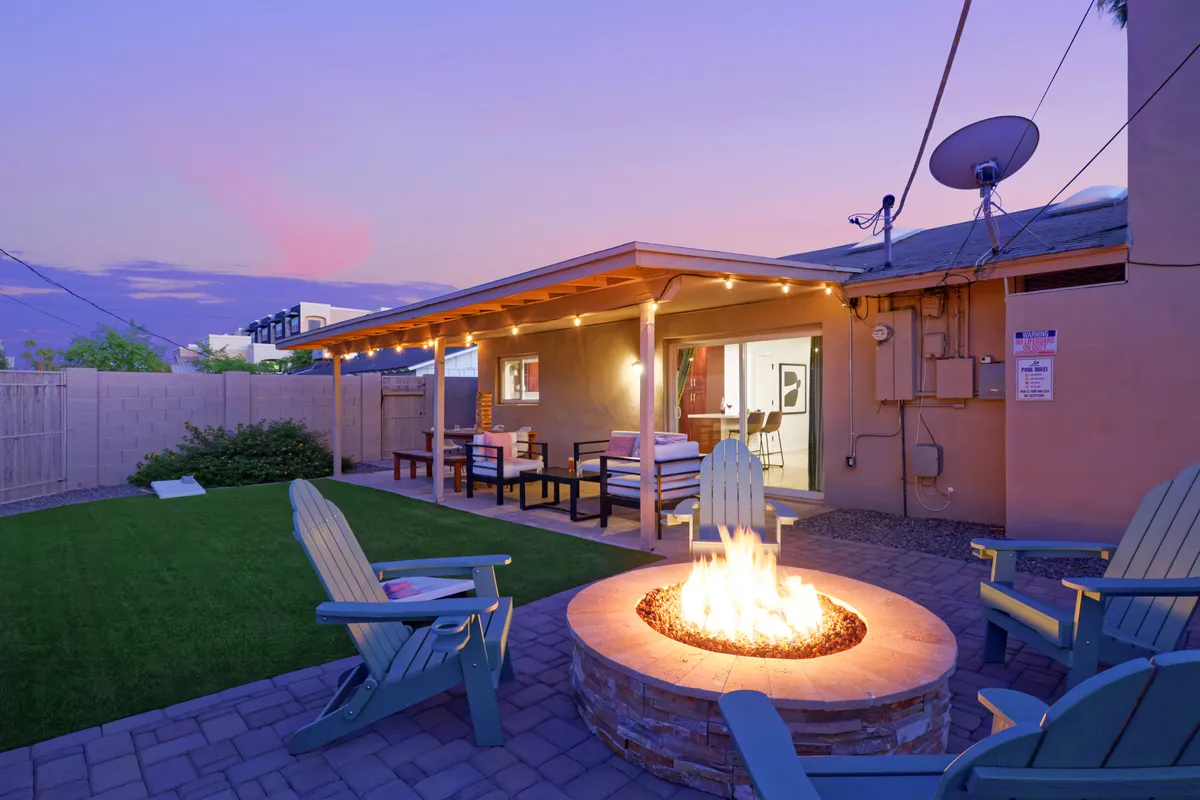 A cozy outdoor seating area centered around a glowing fire pit, illuminated by string lights. The surrounding lawn offers space for yard games, and the atmosphere is warm and inviting, ideal for group gatherings, storytelling, or making s'mores after sunset.