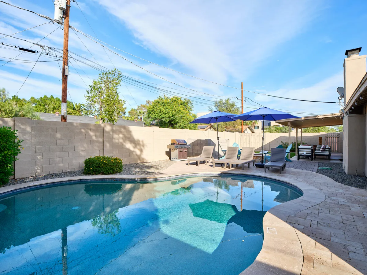 A sun-filled backyard scene featuring a sparkling swimming pool surrounded by comfortable lounge chairs and umbrellas. There's also a grill area, and the lush greenery creates a relaxed, family-friendly setting. The space is clearly designed for both play and relaxation, inviting groups to swim, sunbathe, barbecue, and gather together outdoors.