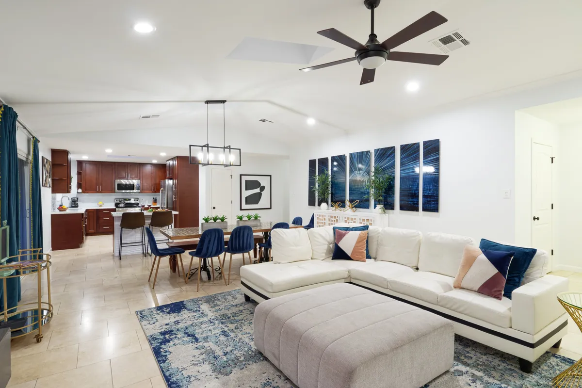 This interior image features a spacious open-concept area with a large sectional sofa, an expansive dining table, wall artwork, and lush decorative plants. The space is filled with natural light, and the arrangement encourages conversation, relaxation, and group activities such as watching TV or dining together.