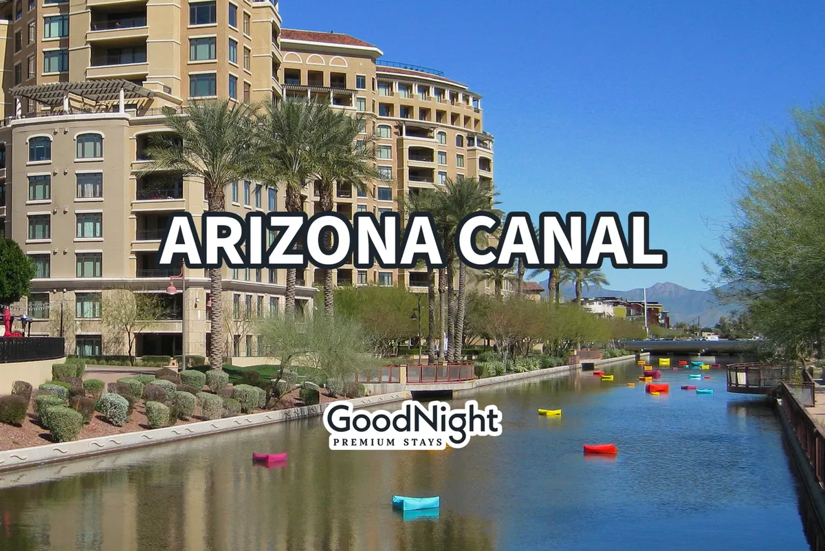 A vibrant view of the Arizona Canal lined with palm trees, colorful boats, and modern buildings. People can be seen strolling along the waterway, enjoying the lively yet relaxed atmosphere. The scene exudes brightness and community, with clear skies and reflections in the water.