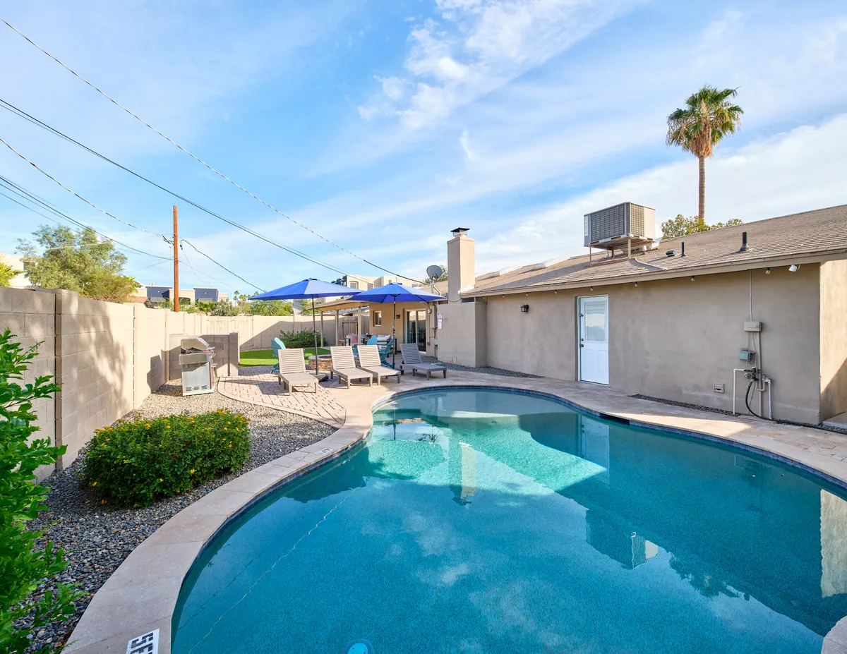 A spacious backyard scene featuring a sparkling pool, patio furniture, outdoor grill, blue umbrellas, and a tall palm tree. The area invites both swimming and outdoor dining, with a seamless blend of leisure and entertainment spaces.