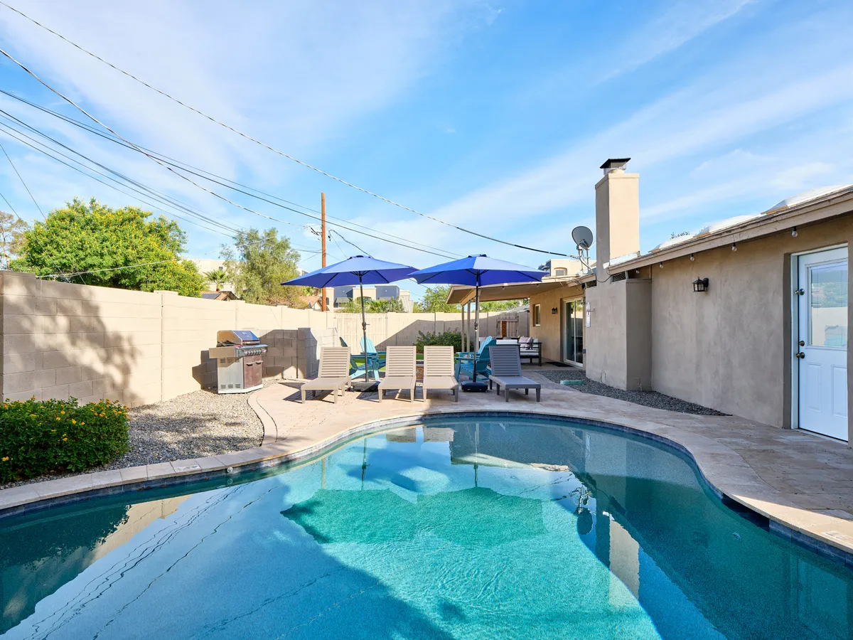 A sparkling swimming pool surrounded by blue umbrellas and inviting lounge chairs, with an outdoor grill nearby. The scene suggests relaxation, social gatherings, and poolside fun in a private resort-style setting.