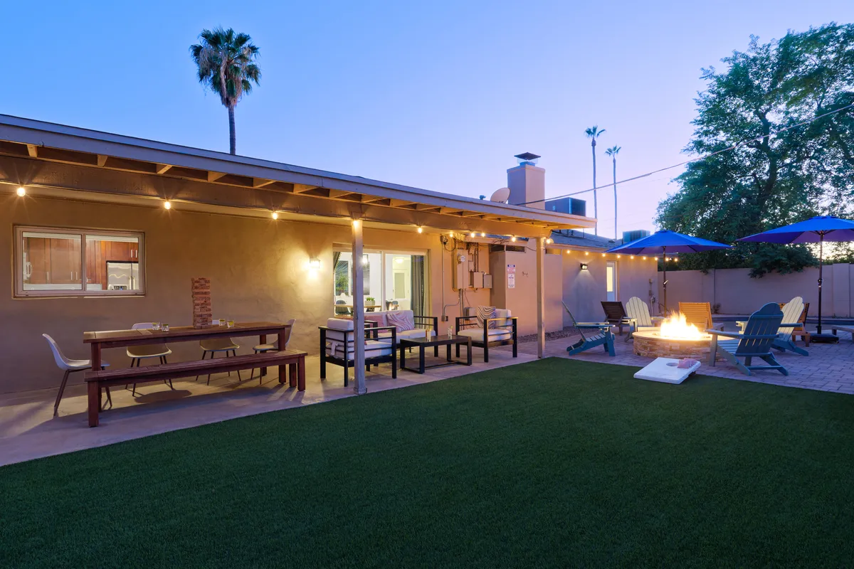 A cozy fire pit area with comfortable outdoor seating, table and chairs, palm tree, umbrellas, and a grassy lawn. The setting is ideal for gathering, sharing stories, or playing yard games as the sun sets.
